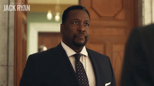 A Man In A Suit And Tie Stands In Front Of A Sign That Says " Jack Ryan " GIF
