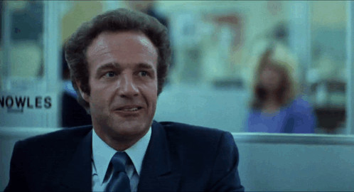 A Man In A Suit And Tie Stands In Front Of A Sign That Says Nowles GIF