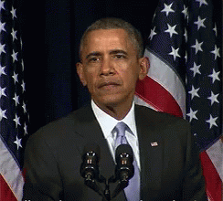 A Man In A Suit And Tie Stands In Front Of Two Microphones In Front Of An American Flag GIF