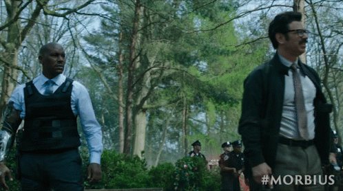 A Man In A Suit And Tie Stands Next To A Man In A Bullet Proof Vest In A Forest GIF