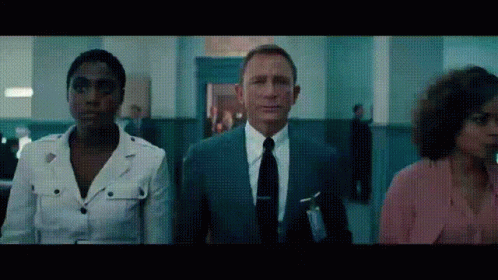 A Man In A Suit And Tie Stands Next To Two Women In A Hallway GIF