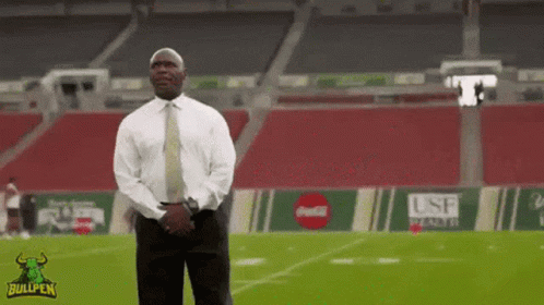 A Man In A Suit And Tie Stands On A Football Field With A Bullpen Logo On The Bottom GIF