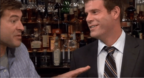 A Man In A Suit And Tie Talks To Another Man In A Bar GIF