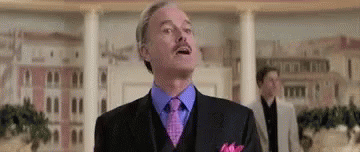 A Man In A Suit And Tie With A Mustache Is Standing In Front Of A Building . GIF