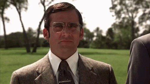 A Man In A Suit And Tie With Glasses Is Standing In A Grassy Field . GIF