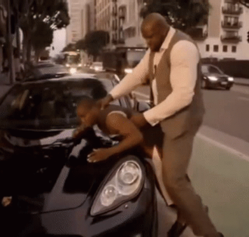 A Man In A Suit And Vest Is Petting Another Man 'S Car GIF