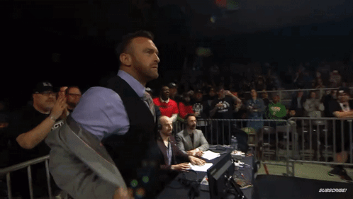 A Man In A Suit And Vest Stands In Front Of A Crowd With A Sign That Says ' Warriors ' On It GIF