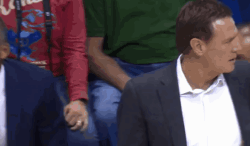 A Man In A Suit And White Shirt Is Sitting In A Stadium With Other Men . GIF