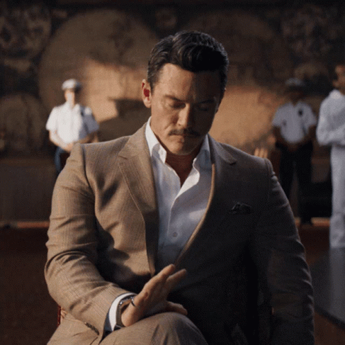 A Man In A Suit And White Shirt Is Sitting In Front Of A Painting GIF