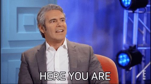 A Man In A Suit And White Shirt Is Sitting In Front Of A Sign That Says Here You Are GIF