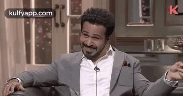 A Man In A Suit And White Shirt Is Sitting On A Couch With His Arms Outstretched And Smiling . GIF
