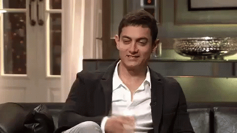 A Man In A Suit And White Shirt Is Sitting On A Couch With His Legs Crossed And Making A Funny Face . GIF