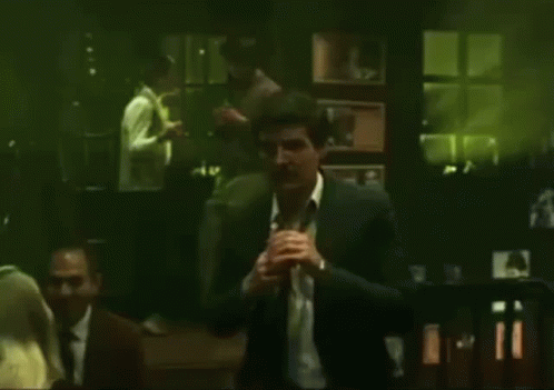 A Man In A Suit And White Shirt Is Standing In A Dark Room With A Green Background . GIF