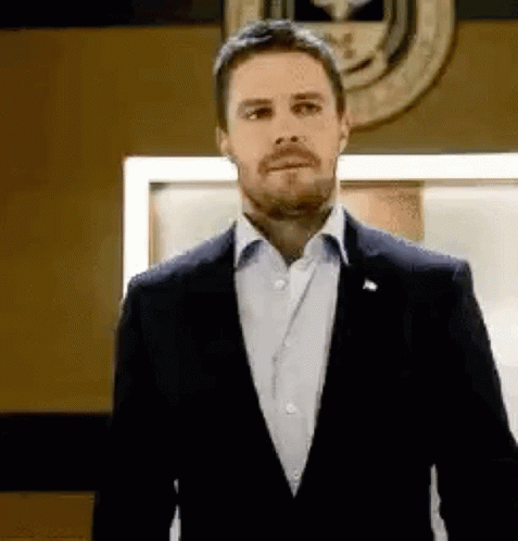 A Man In A Suit And White Shirt Is Standing In Front Of A Wall With A Seal On It . GIF