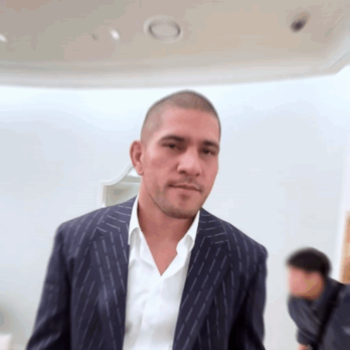 A Man In A Suit And White Shirt Stands In Front Of A White Wall GIF