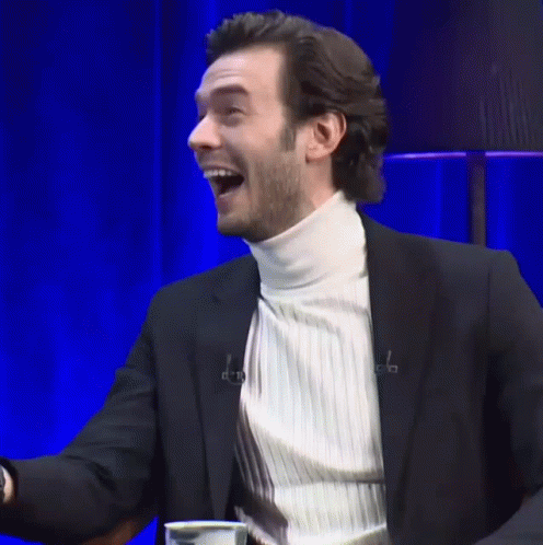 A Man In A Suit And White Turtleneck Is Laughing With His Mouth Open GIF