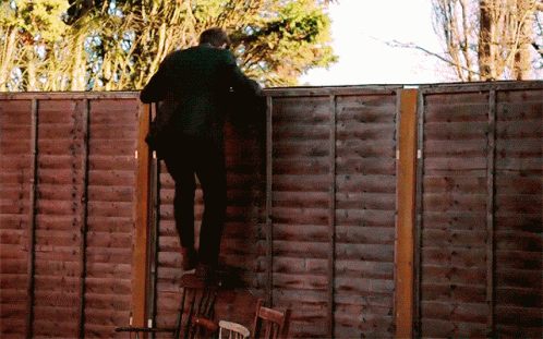 A Man In A Suit Climbs A Wooden Fence With Trees In The Background GIF