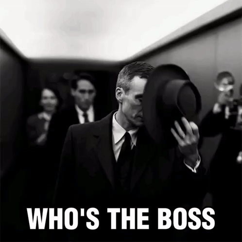 A Man In A Suit Covering His Face With His Hat And The Words Who 'S The Boss Behind Him GIF