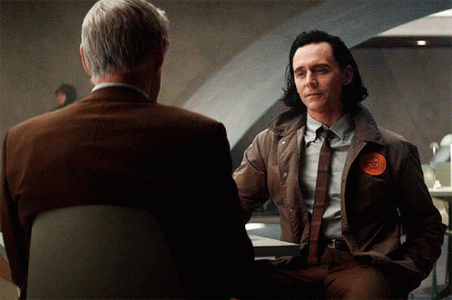 A Man In A Suit Has A Patch On His Jacket That Says ' Loki ' GIF