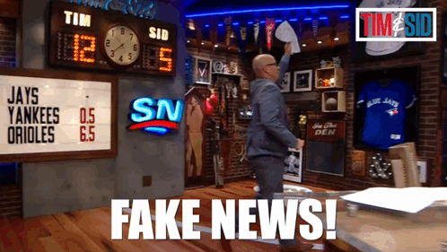 A Man In A Suit Holds A Piece Of Paper In Front Of A Scoreboard That Says Fake News GIF