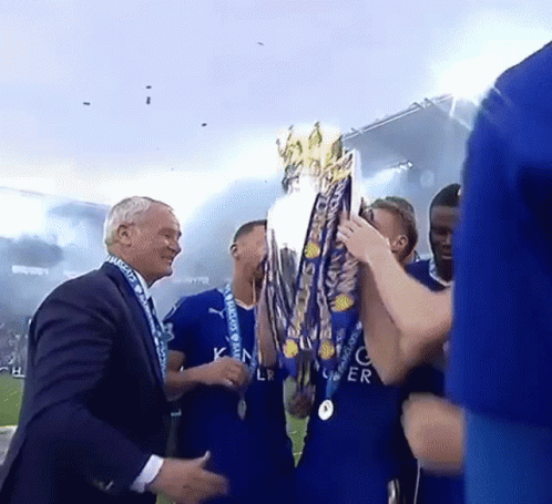 A Man In A Suit Holds A Trophy In Front Of A Group Of Soccer Players In Blue Jerseys GIF