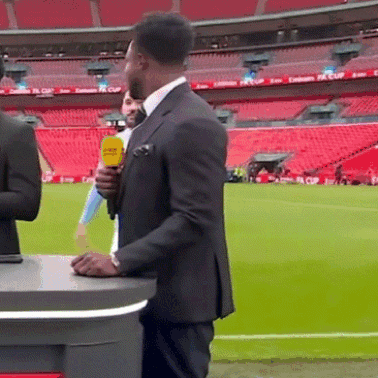 A Man In A Suit Holds A Yellow Microphone In Front Of A Stadium That Has Emirates Fa Cup Written On The Seats GIF