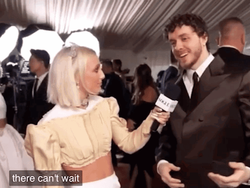 A Man In A Suit Is Being Interviewed By A Woman Who Is Holding A Microphone That Says Vogue GIF