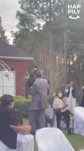 A Man In A Suit Is Carrying A Baby In His Arms While A Group Of People Are Sitting In Chairs . GIF