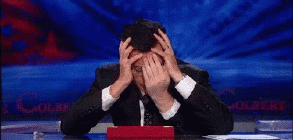 A Man In A Suit Is Covering His Face With His Hands While Sitting At A Desk GIF