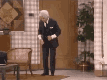 A Man In A Suit Is Dancing In A Living Room With A Chair And A Plant . GIF