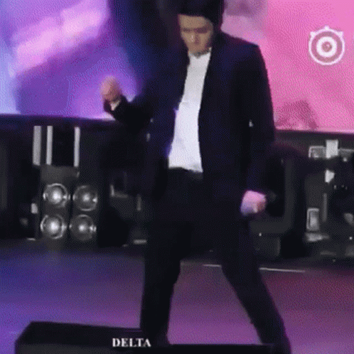 A Man In A Suit Is Dancing On A Stage In Front Of Delta Speakers GIF