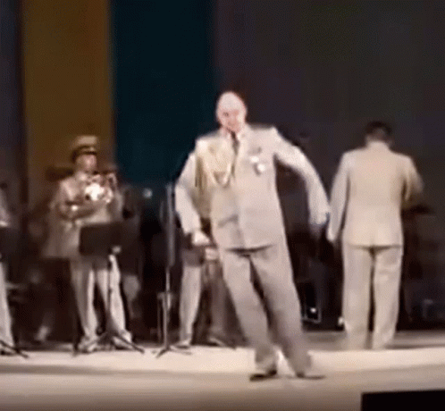 A Man In A Suit Is Dancing On A Stage While A Band Plays Behind Him . GIF