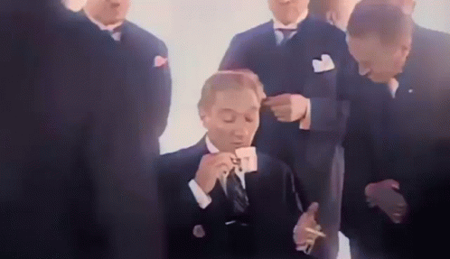A Man In A Suit Is Drinking A Cup Of Coffee And Smoking A Cigarette While A Group Of Men Look On . GIF