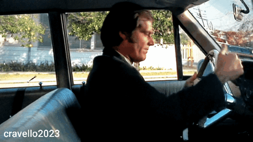 A Man In A Suit Is Driving A Car With Cravello2023 Written On The Bottom Right GIF
