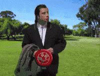 A Man In A Suit Is Holding A Frisbee In A Park With Trees In The Background GIF