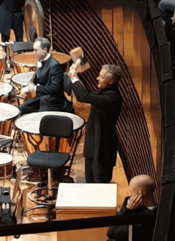A Man In A Suit Is Holding A Hammer In His Hand In Front Of A Drum Set GIF