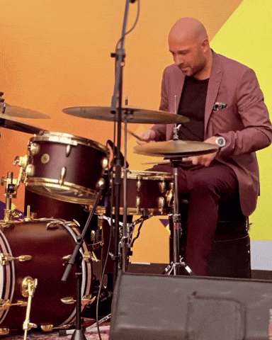 A Man In A Suit Is Playing Drums In Front Of A Yellow Wall GIF