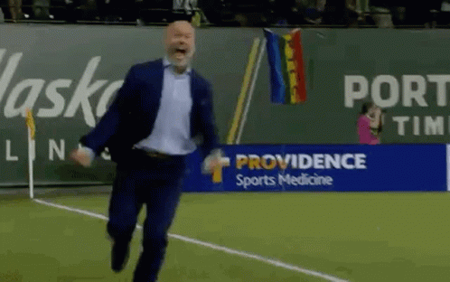 A Man In A Suit Is Running On A Soccer Field In Front Of A Providence Sports Medicine Banner . GIF