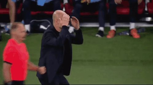 A Man In A Suit Is Running On A Soccer Field With A Referee In A Red Shirt Behind Him GIF
