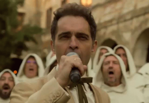 A Man In A Suit Is Singing Into A Microphone In Front Of A Group Of Monks In White Robes . GIF