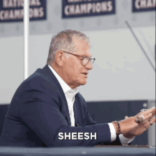 A Man In A Suit Is Sitting At A Table And The Word Sheesh Is On His Arm GIF