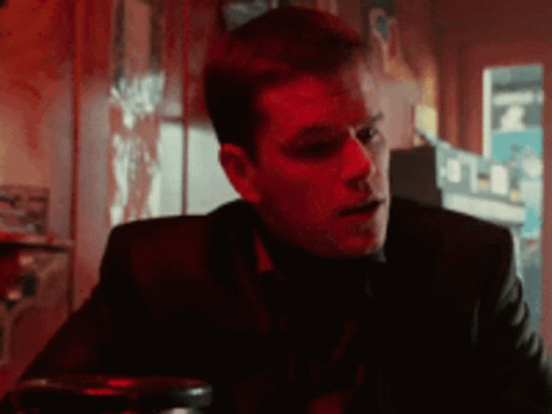 A Man In A Suit Is Sitting At A Table In A Dark Room With A Red Light Behind Him . GIF