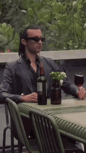 A Man In A Suit Is Sitting At A Table With A Bottle Of Wine And A Glass . GIF