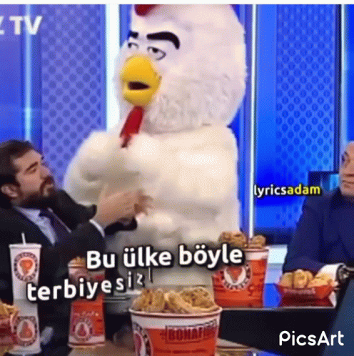 A Man In A Suit Is Sitting At A Table With A Chicken Mascot Standing Next To Him . GIF