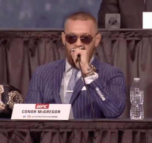 A Man In A Suit Is Sitting At A Table With A Sign That Says Conor Mcgregor On It GIF