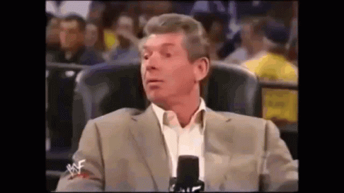 A Man In A Suit Is Sitting In Front Of A Microphone In A Crowd . GIF
