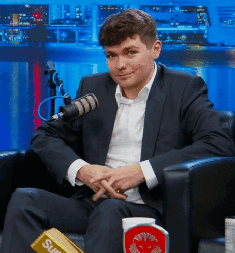 A Man In A Suit Is Sitting In Front Of A Microphone With A Supreme Bar On His Lap GIF