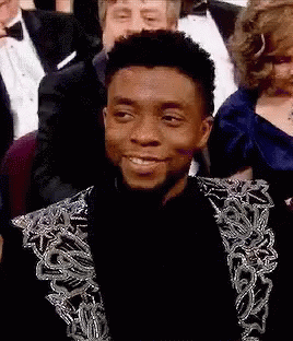 A Man In A Suit Is Smiling While Sitting In A Crowd . GIF