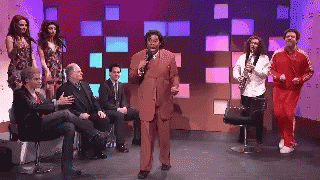A Man In A Suit Is Speaking Into A Microphone While A Group Of People Sit Around Him GIF