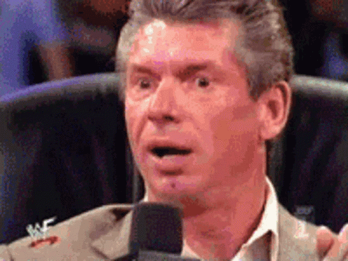 A Man In A Suit Is Speaking Into A Microphone With A Wrestler Logo On His Jacket GIF